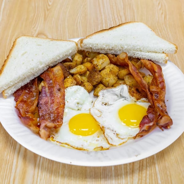 A breakfast plate with two fried eggs, crispy bacon, seasoned home fries, and white toast.