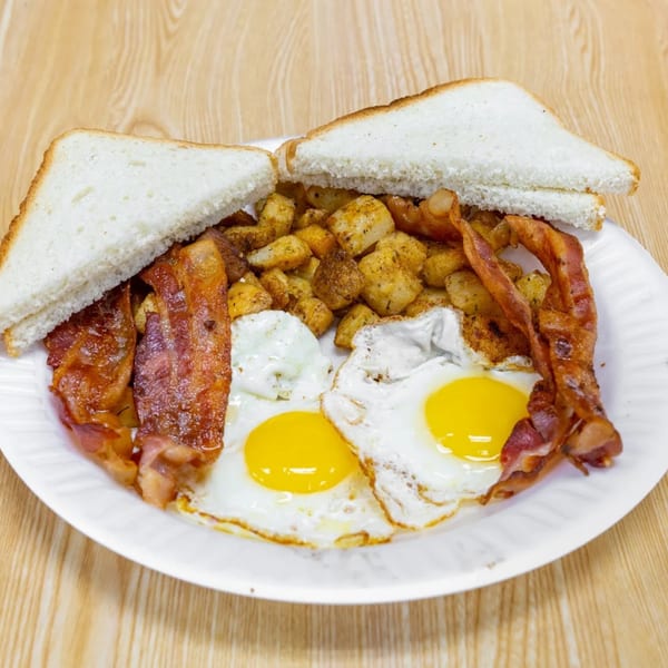 A breakfast plate with two fried eggs, crispy bacon, seasoned home fries, and white toast.