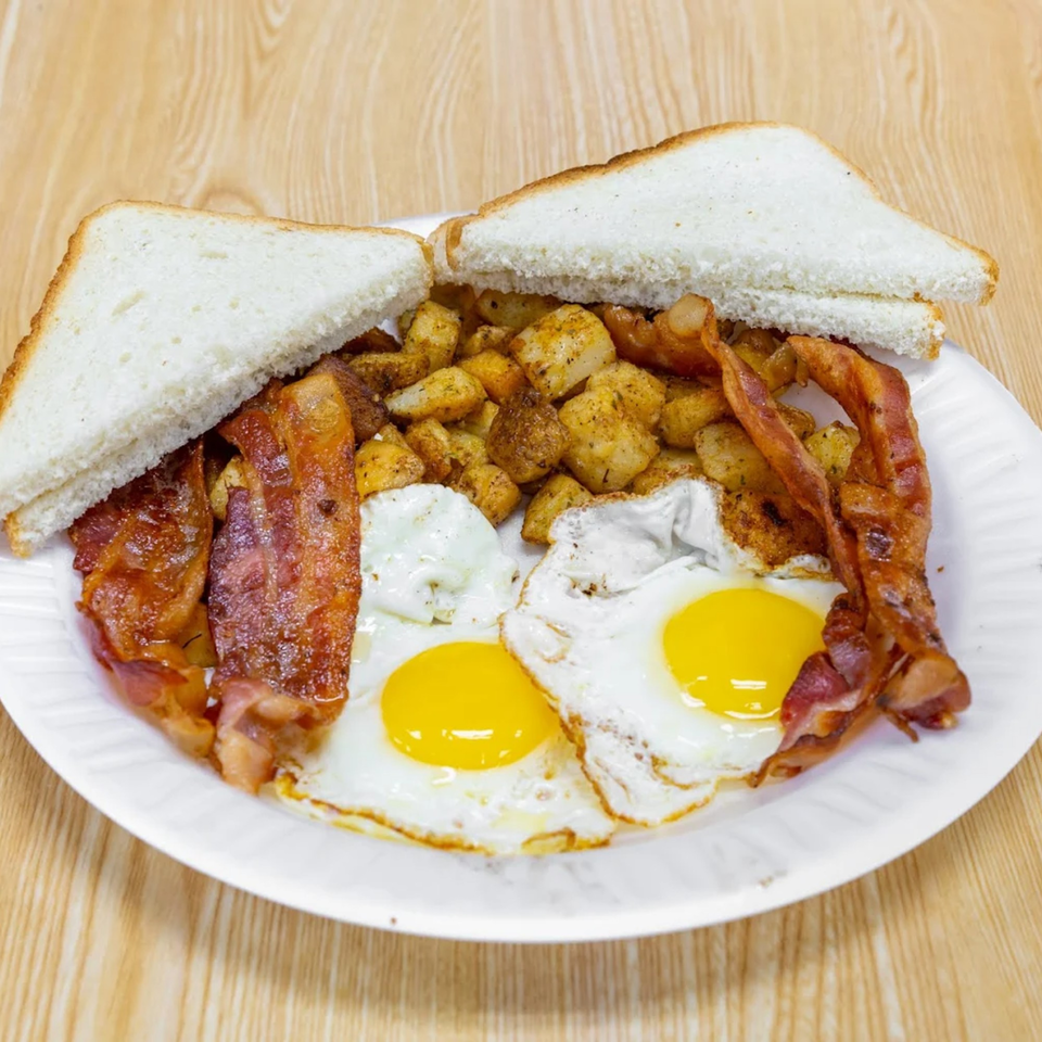 A breakfast plate with two fried eggs, crispy bacon, seasoned home fries, and white toast.