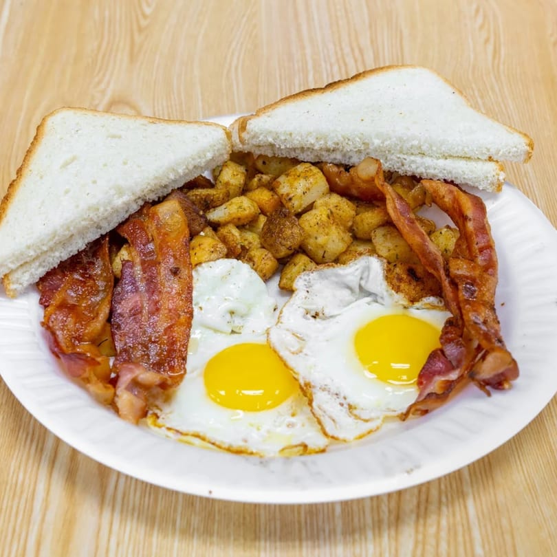 A breakfast plate with two fried eggs, crispy bacon, seasoned home fries, and white toast.