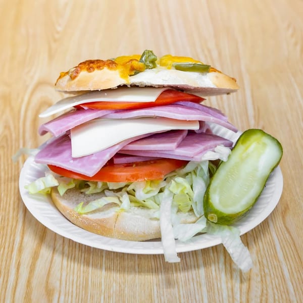 A large deli sandwich with ham, cheese, lettuce, and tomatoes, served with a pickle spear.