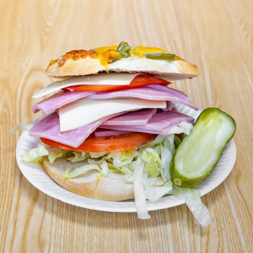 A large deli sandwich with ham, cheese, lettuce, and tomatoes, served with a pickle spear.