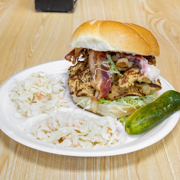Grilled chicken sandwich with bacon, lettuce, coleslaw, and a pickle on a white plate.